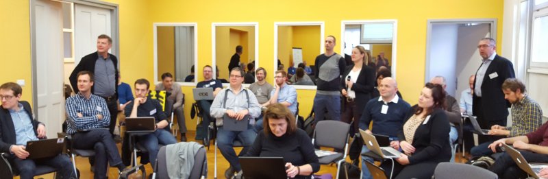 A seminar room with several people sitting and standing. Centre of the picture, a woman holds a microphone